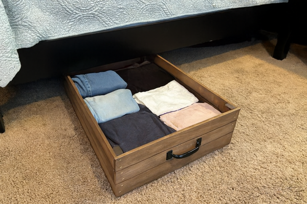 Rustic wooden under-bed storage drawer filled with folded jeans, shirts, and casual clothes for organized bedroom storage.