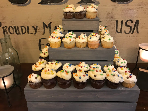 Close-up of 3-tier wooden cupcake stand in antique gray stain filled with cupcakes with candy toppings.