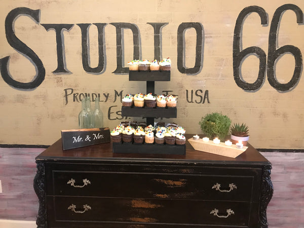 Rustic three-tier dark cupcake stand loaded with cupcakes against a Studio 66 event backdrop.