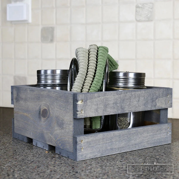 Small rustic gray crate used in kitchen for storage with jars and towels.