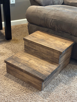 Angled view of a rustic medium brown 2-step stool on carpet in front of a recliner.