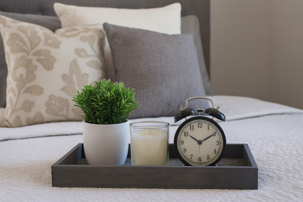 Small gray wooden tray on bed with plant, candle, and alarm clock decor.