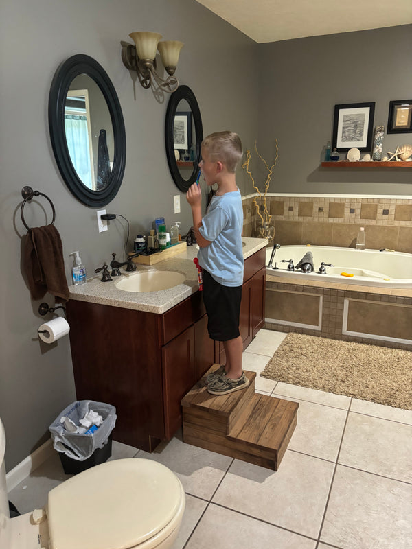 Child standing on 2-tier Early American pet steps while brushing teeth at bathroom sink.